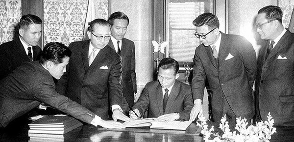 1965년 12월 18일 한일 협정 비준서에 서명하는 박정희(앉은 이). 한일 관계 정상화가 한국의 베트남 참전을 가능케 했다. 미국은 한일 협정으로 한국이 정상화됨에 따라 베트남에서 미국과 동맹관계를 맺을 수 있다고 판단했다. (국가기록원)