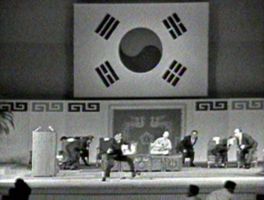 1974년 8월 15일 국립극장 대극장에서 일어난 대통령 부인 육영수 피격 장면. 경호실장 박종규가 권총을 뽑아들고 단상 앞으로 뛰쳐나온 가운데 뒤편 의자에 앉은 한복 차림의 육영수가 총탄에 맞아 옆으로 쓰러지고 있다.