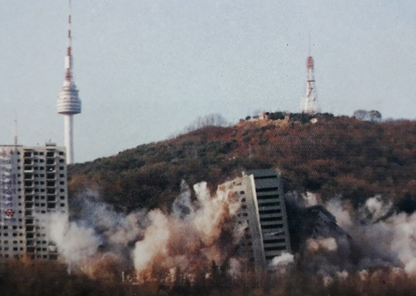 1994년 11월 20일 오후 3시, 남산 외인아파트 건물이 발파 해체 공법으로 철거되는 장면이 TV로 중계되고 있다.